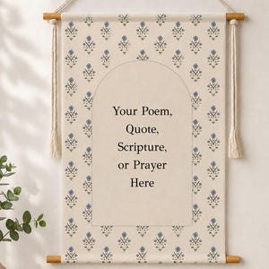 May include: A cream-coloured fabric wall hanging suspended from two wooden dowels with a white rope. The fabric features a repeating blue floral pattern and a central arch outline containing the text: Your Poem, Quote, Scripture, or Prayer Here.