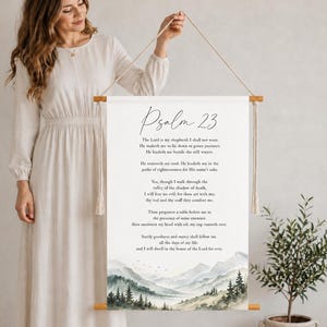 May include: A white fabric wall hanging featuring the text of Psalm 23 in black script and serif fonts. The bottom of the banner displays a watercolour landscape of mountains and pine trees. It is suspended by two wooden rods and a beige cord.