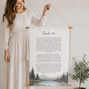 Könnte beinhalten: Ein weißer Wandbehang aus Stoff mit dem Text von Psalm 42 in schwarzer Schrift. Am unteren Rand der Bannereinheit befindet sich eine Aquarelllandschaft mit See, Wald und Bergen. Er ist an zwei Holzstäben und einer dünnen, hellen Schnur aufgehängt.