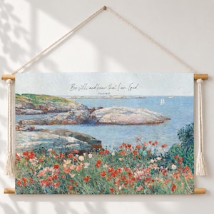 May include: A rectangular wall hanging featuring a landscape painting of a rocky coastline with red flowers in the foreground and the text Be still, and know that I am God. Psalm 46:10. The canvas is suspended from a wooden rod with white rope.