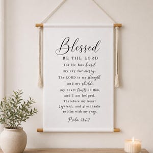 May include: A white fabric wall hanging featuring black printed text that reads: Blessed BE THE LORD for He has heard my cry for mercy. The LORD is my strength and my shield; my heart trusts in Him, and I am helped. Psalm 28:6-7.