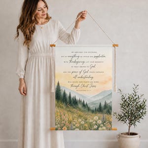 Könnte beinhalten: Ein Wandbehang aus Stoff mit einer Aquarell-Landschaft aus einer Blumenwiese und einem Kiefernwald unter einem sanften orangefarbenen Himmel. Der Text lautet: Be anxious for nothing, but in everything by prayer and supplication, with thanksgiving, let your requests be made known to God; and the peace of God, which surpasses all understanding, will guard your hearts and minds through Christ Jesus. Philippians 4:6-7.
