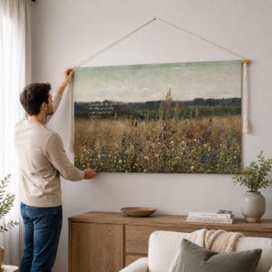 May include: A rectangular wall hanging featuring a landscape painting of a wildflower field with blue and white flowers under a pale sky. The artwork includes the text "Consider the lilies of the field, how they grow; they neither toil nor spin."