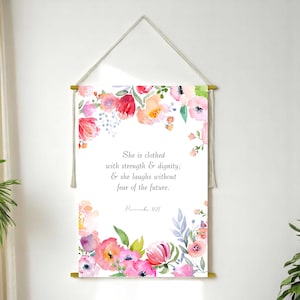 Könnte beinhalten: Ein floraler Wandbehang mit weißem Hintergrund und goldfarbenen Holzdübeln. Das Kunstwerk zeigt ein Aquarell-Blumenmuster in Rosa-, Rot- und Orangetönen. Der Text lautet: "She is clothed with strength & dignity; & she laughs without fear of the future."