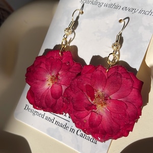 May include: Earrings with pressed red rose petals, suspended from gold-coloured hooks. The earrings are displayed on a white card with text: "Inspired by nature, sparkling within every inch. Designed and made in Canada."