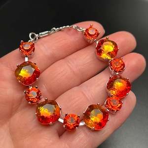 May include: A silver bracelet featuring alternating large and small orange and yellow gemstones. The gemstones are faceted and set in silver prongs, creating a sparkling and eye-catching accessory. The bracelet is shown against a dark background.