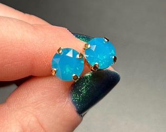 May include: A pair of round, light blue stud earrings with gold-coloured settings. The earrings have a faceted appearance, catching the light. The background is a dark, blurred colour.