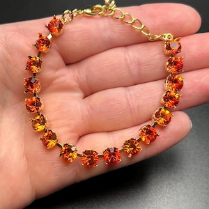 Peut inclure: Un bracelet doré orné de pierres précieuses orange-rouge. Les pierres rondes sont serties sur une chaîne délicate. Le bracelet est présenté sur un fond neutre, mettant en valeur les couleurs chaudes du bijou.