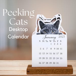 May include: A desktop calendar featuring a gray cat peeking over the top. The calendar displays the month of January 2025, with the days of the week and dates clearly visible. The calendar is set on a wooden base, with the words "Peeking Cats Desktop Calendar" displayed.