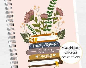 Puede incluir: Un diseño floral rosa y blanco con una pila de libros que dice "Slow progress is still progress". El texto "Your name here Lesson Planner" está en la parte inferior de la imagen. El texto "Available in 6 different cover colours" está en un círculo en el lado derecho de la imagen.