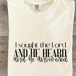 Puede incluir: Camiseta Comfort Colors color crema con el texto negro "I sought the Lord AND HE HEARD and he answered". La camiseta tiene cuello redondo y una etiqueta en el cuello.