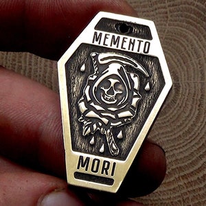 Może przedstawiać: Brązowy wisiorek w kształcie trumny z czarno-białym obrazkiem Kosy trzymającej kosę i różę. Na wisiorku wygrawerowano tekst "MEMENTO MORI".