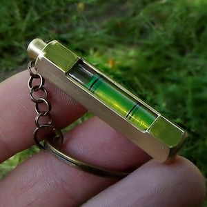 May include: A small, brass-colored level with a green liquid bubble, attached to a keychain. The level is rectangular with a hexagonal end. The bubble is used to determine if a surface is level. The background is blurred green.