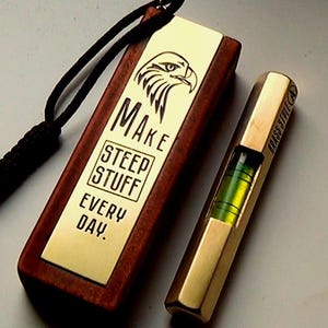 May include: A wooden keychain with a brass plate engraved with the text "MAKE STEEP STUFF EVERY DAY." and an eagle design. The keychain is attached to a black cord. A brass level with a green bubble is also included.