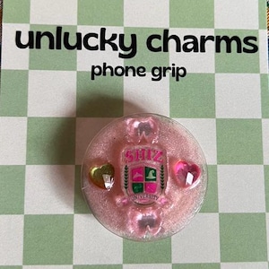 Puede incluir: Un soporte para teléfono rosa con un escudo universitario y detalles en forma de corazón, exhibido en una tarjeta con el texto "unlucky charms phone grip" sobre un patrón de cuadros verdes y blancos.