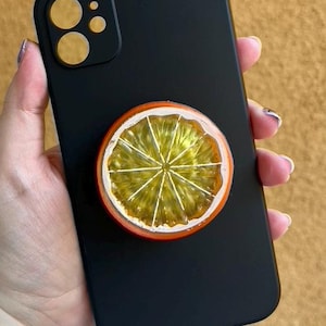 May include: Black smartphone case with a vibrant orange slice pop-up grip. The orange slice features a detailed design with yellow segments and a red rind. The phone case has a matte finish and a camera bump with three lenses.