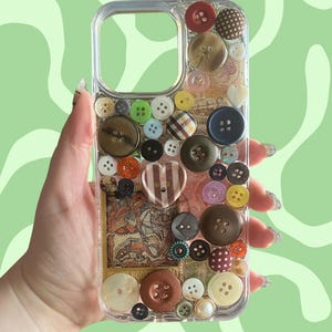 May include: Clear phone case decorated with an assortment of colorful buttons in various sizes and styles. The case features a heart-shaped button and a vintage postage stamp design. The background is a light green wavy pattern.
