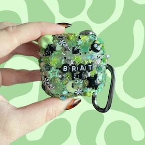 May include: A lime green protective case for wireless earbuds, heavily decorated with various charms including black hearts, stars, small flowers, and a cartoon character. The word BRAT is spelled out in black and white letter beads across the center.