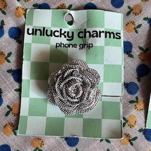 May include: A silver rose-shaped phone grip on a green and white checkered card. The card reads "unlucky charms phone grip". The background is a fabric with a pattern of blue and yellow fruit.