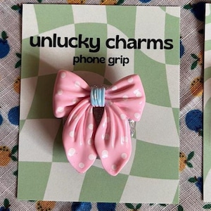 May include: A pink bow-shaped phone grip with white polka dots and a light blue center. The phone grip is on a green and white checkered card that reads "unlucky charms phone grip."