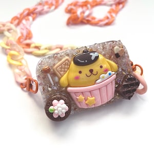 May include: A yellow dog character wearing a blue beret sits in a pink cupcake with a brown waffle, a honey stick, and a chocolate flower. The cupcake is surrounded by glitter and small white crystals. The dog character has a happy expression and is wearing a pink and white striped shirt. The cupcake is decorated with two yellow stars. The necklace is made of a multi-colored chain.