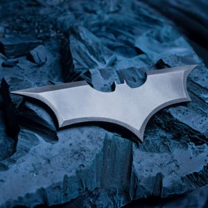 May include: A silver-colored Batman logo emblem with sharp, angular edges. The emblem is set against a textured, dark blue background, creating a contrast. The iconic bat-shaped symbol is a recognizable design.