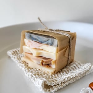 May include: A stack of four rectangular soap bars wrapped in brown paper and tied with twine, resting on a small cream-colored crocheted cotton pouch. The top soap bar is marbled gray and white, while the others are shades of cream and peach.