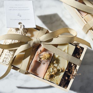 May include: A handmade soap set in a wooden box, tied with a beige ribbon. The box contains several bars of soap with natural ingredients. A tag with the text "SHEADOW SOAP CO." is attached to the box.