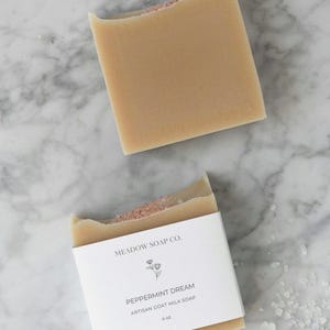 May include: Two rectangular bars of artisan goat milk soap. One bar is whole, the other has a bite taken out of it. The bottom bar has a white label that reads "Meadow Soap Co." and "Peppermint Dream" with a floral graphic. The soap is 4 oz.