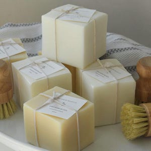 May include: A collection of square, cream-colored soaps tied with twine and labeled, alongside two wooden handled scrub brushes with yellow bristles. The items are arranged in a white, oval dish with a gray and white striped cloth.