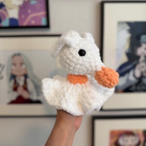 May include: A white crocheted plush toy shaped like a ghost with a rounded head, floppy ears, and a ruffled base. The ghost features a bright orange collar and a matching orange nose, with a single visible black plastic eye on the side of its head.