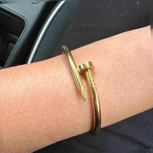 May include: A gold-toned metal bangle bracelet shaped like a bent nail. The bracelet features a smooth, curved shank that tapers to a point at one end and terminates in a flat, cylindrical nail head with etched horizontal lines at the other end.