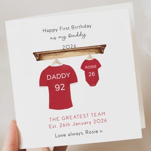 May include: A white birthday card with the text "Happy First Birthday as my Daddy 2026." It shows a red jersey with "DADDY 92" and a red babygrow with "ROSIE 26" hanging from a wooden shelf. The text "THE GREATEST TEAM" is also present.