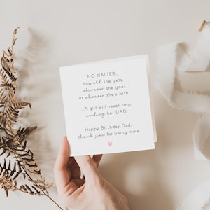May include: A white greeting card with black text, held by a hand, reads "Happy Birthday Dad." A dried fern and white ribbon are arranged on a neutral background. A small pink heart is printed on the card.