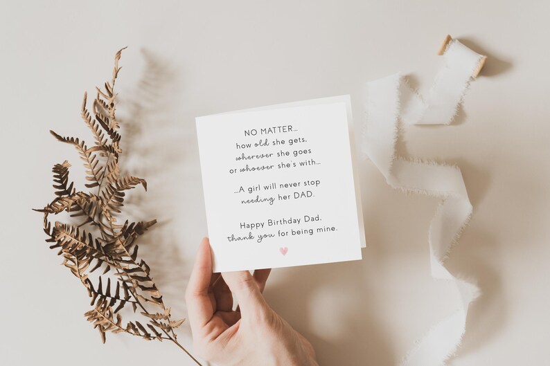 May include: A white greeting card with black text, held by a hand, reads "Happy Birthday Dad." A dried fern and white ribbon are arranged on a neutral background. A small pink heart is printed on the card.