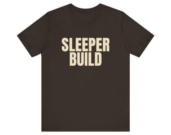 May include: A dark brown short-sleeved t-shirt featuring the text SLEEPER BUILD printed in a large, bold, cream-coloured sans-serif font across the centre of the chest. The garment has a classic crew neckline and a standard straight-cut hem.