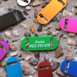 May include: Assortment of colorful metal dog tags with engraved text. The tags are in various colors, including green, orange, pink, blue, red, black, and white. One tag has the text "Jessie 0412 345 678" engraved on it.