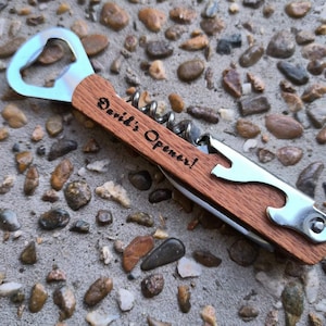 Puede incluir: Un abridor de botellas multiusos con mango de madera. El mango está grabado con el texto "David's Opener!"