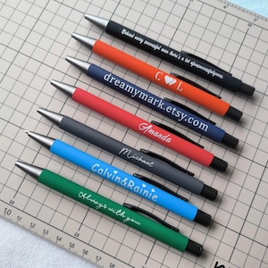 May include: Eight personalised pens with different coloured bodies and white text. The text includes names, a quote, and a website address.