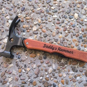 May include: A claw hammer with a black head and a wooden handle. The handle is engraved with the words "Daddy's Hammer". The hammer is lying on a stone surface.