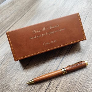 Puede incluir: Un estuche de cuero marrón y un bolígrafo de madera con detalles dorados. El estuche tiene grabado el texto "Dear Ms. Amanda, thank you for helping me grow. Colin 2025". El bolígrafo está grabado con el nombre "Amanda".