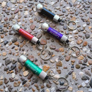 Może przedstawiać: Cztery latarki do kluczy z różnokolorowymi korpusami i srebrnymi kółkami do kluczy. Latarki mają nadrukowane współrzędne: 27.4705° S 153.0260° E, 27.4705° S 153.0260° E i "Manuka".