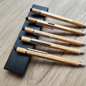 Puede incluir: Cinco bolígrafos de bambú con detalles plateados y puntas de lápiz táctil negras se muestran en una caja negra. Los bolígrafos tienen un diseño de regla y nivel. Un bolígrafo está grabado con el texto "Dear Mr. James, Thank you for helping me grow. Colin 2025."
