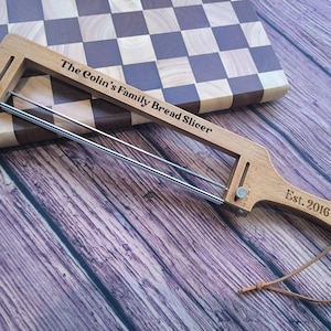 May include: A wooden bread slicer with a metal wire cutting mechanism, featuring the engraved text "The Colin's Family Bread Slicer" and "Est. 2016". The tool includes a handle with a leather hanging cord and an adjustable metal guide.