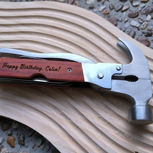 May include: A multi-tool hammer with a wooden handle, featuring a hammer head, pliers, and a knife. The handle is engraved with the text "Happy Birthday, Colin!" The tool is silver and brown, resting on a carved wooden surface.