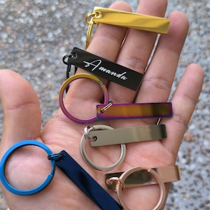 May include: A set of six keychains with different coloured metal tags. The tags are rectangular and have a smooth, polished finish. One tag is black with the name "Amanda" engraved on it. The other tags are gold, silver, blue, rainbow, and rose gold.