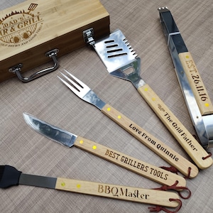 May include: A wooden barbecue tool set with a bamboo box. The set includes a spatula, fork, knife, brush, and tongs. The tools have wooden handles with engraved text, including "Dad's Bar & Grill" and "BBQ Master."