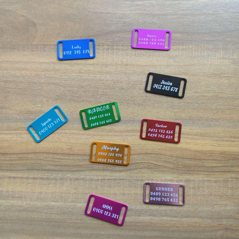 May include: Assortment of colorful metal dog tags with engraved names and phone numbers. The tags are in various colors, including pink, red, orange, green, blue, and purple.