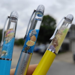 May include: Three pens with clear barrels filled with blue, yellow, and clear liquid, and miniature seashells. The pens have silver clips and tips. The blue pen has the word "uation!" printed on it.