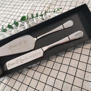 以下が含まれることがあります： 黒い箱に入った銀製のケーキナイフとサーバーのセット。ナイフとサーバーには「Colin & Lin 26.11.2024」という文字が刻まれています。